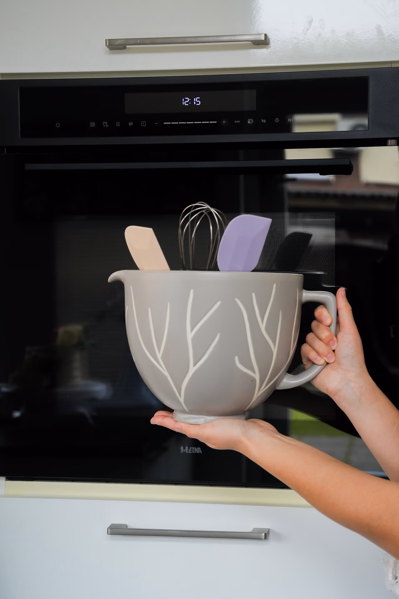 Vera van Baksels van Vera houdt een grijze keramische mengkom met bakspatels en een garde vast in een moderne keuken.