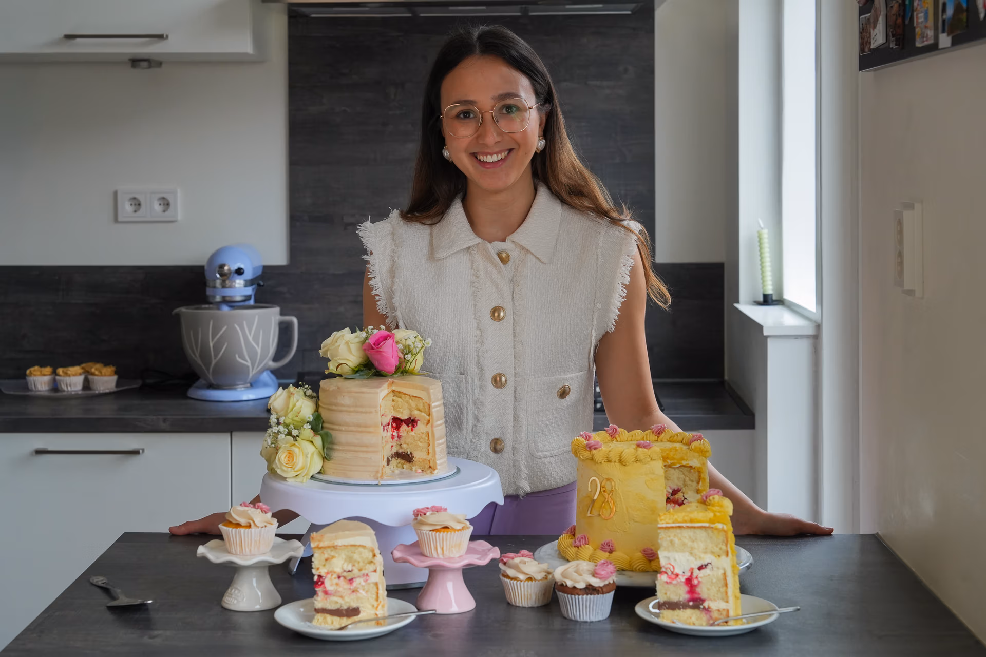 Aanrecht met twee aangesneden taarten en meerdere cupcakes, met Vera, eigenaar van Baksels van Vera in een moderne keuken op de achtergrond.