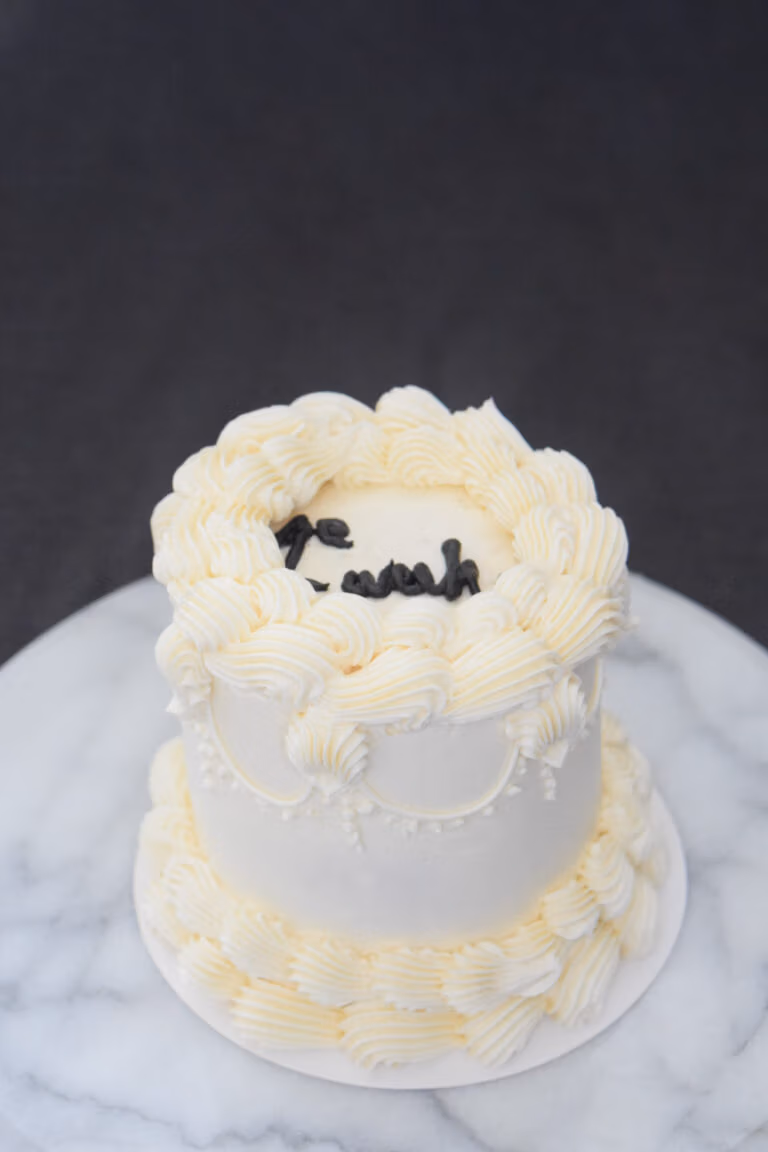 Witte ronde vintage bento taart met elegante frosting‑swirls en zwarte tekst op marmeren ondergrond.