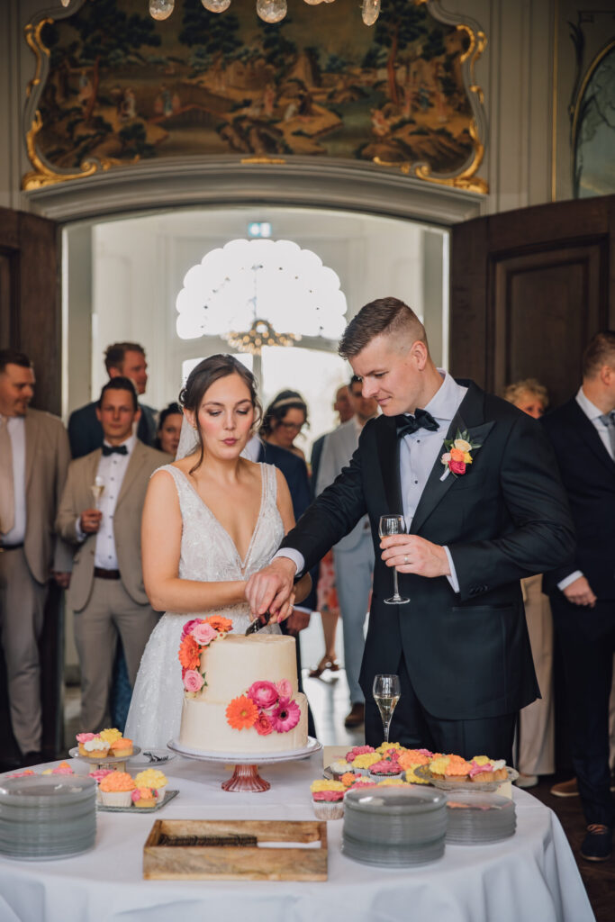 Bruidspaar snijdt de witte bruidstaart aan. Bruidstaart met roze en oranje bloemen.