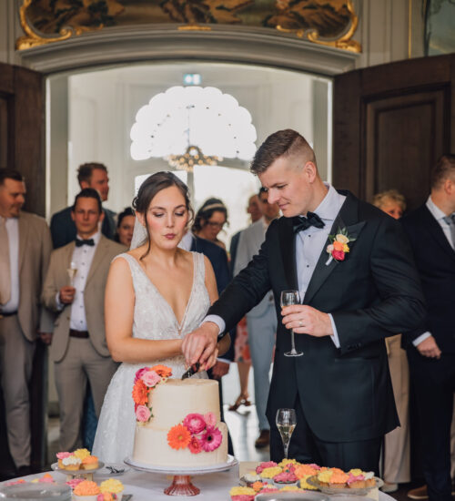 Bruidspaar snijdt de witte bruidstaart aan. Bruidstaart met roze en oranje bloemen.